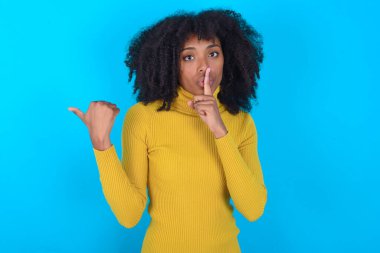 Mavi arka planda sarı boğazlı kazak giyen Afro-Amerikalı bir kadın ellerini yana doğru uzatarak parmaklarını dudaklarına dayayıp sessiz olmak istiyor. Sessizlik ve gizli konsept.
