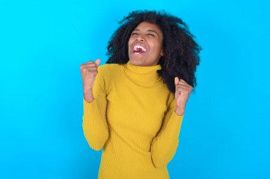 Mavi arka plan üzerine sarı balıkçı yaka giyen Afro-Amerikalı kadın kolları havada ve gözleri kapalı bir şekilde başarıyı kutluyor ve şaşırıyor. Kazanan konsept.