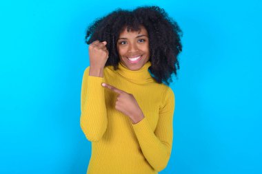 Mavi arka planda sarı boğazlı kazak giyen Afro-Amerikalı kadın. Aceleyle kol saatini işaret ediyor, sabırsızlıkla kameraya bakıyor.