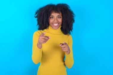 Mavi arka planda sarı boğazlı kazak giyen Afro-Amerikalı kadın mutlu ve komik yüzlü parmaklarını kameraya doğrultuyor. İyi enerji ve titreşimler.