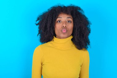 Mavi arka planda sarı boğazlı kazak giyen, dudaklarını büzen, kameraya bakan, insan yüz ifadeleri takınan Afro-Amerikan kadın.