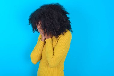 Mavi arka planda sarı balıkçı yaka giyen üzgün Afro-Amerikalı kadın elleriyle yüzünü kapatarak ağlıyor..