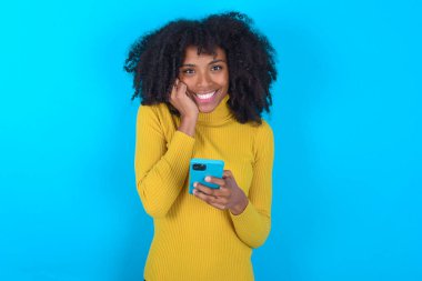 Mavi arka plan üzerine sarı boğazlı kazak giyen neşeli Afro-Amerikalı kadın cep telefonu cihazıyla poz veriyor, modern akıllı telefona mesaj yazıyor, boş zamanlarında komik videolar izliyor, iyi internet bağlantılarından hoşlanıyor,