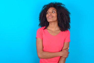 Mavi arka planda pembe tişört giyen mutlu Afro-Amerikalı kadın ellerini göğsünün üzerinden uzatıyor.