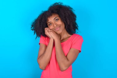 Mavi arka planda pembe tişört giyen çekici Afro-Amerikan kadın kameraya nazikçe gülümsüyor.