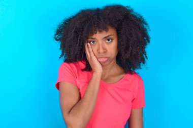 Mavi arka planda pembe tişört giyen üzgün yalnız Afrikalı Amerikalı kadın yanaklarına dokunuyor, dudaklarını ısırıyor ve hoşnutsuzca bakıyor. Kötü duygular