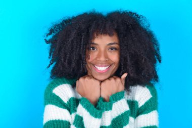 Mavi arka planda çizgili süveter giyen, tatmin olmuş Afro-Amerikalı kadın çeneye iki eliyle dokunuyor, güzel gülümsüyor, sevgilisiyle mutlu bir gün geçiriyor.