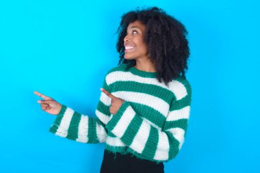 İyimser Afro-Amerikan kadın çizgili süveter giyiyor mavi arka planda iki eliyle ve boş alana bakıyor.