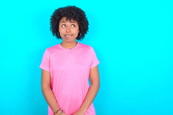 Amazed Young African American woman wearing pink T-shirt over blue background bitting lip and looking tricky to empty space.