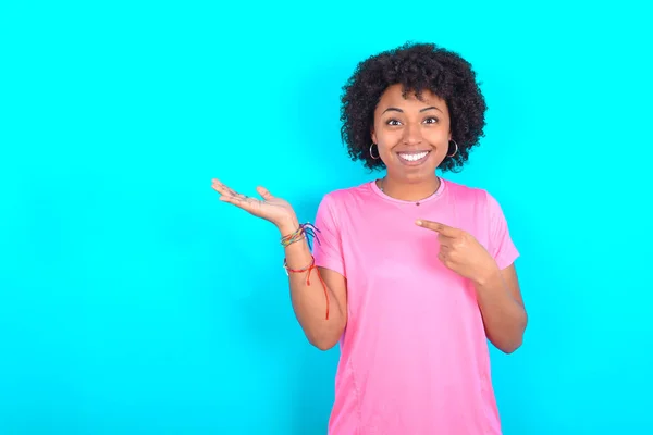 Young African American woman wearing pink T-shirt over blue background pointing and holding hand showing adverts