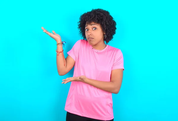 Young African American woman wearing pink T-shirt over blue background pointing aside with both hands showing something strange and saying: I don\'t know what is this. Advertisement concept.