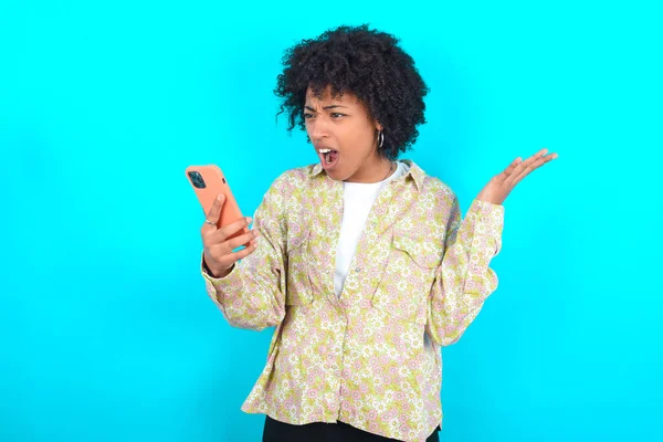 Angry Young African American woman wearing floral shirt over blue background screaming on the phone, having an argument with an employee. Troubles at work.