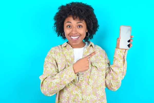 Attractive cheerful Young African American woman wearing floral shirt over blue background holding in hands cell showing black screen