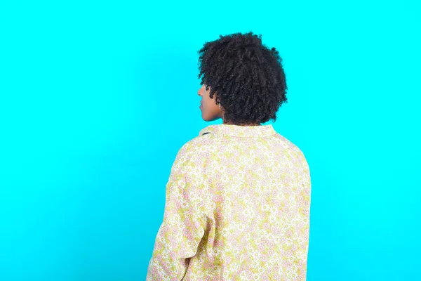 The back side view of a Young African American woman wearing floral shirt over blue background . Studio Shoot.
