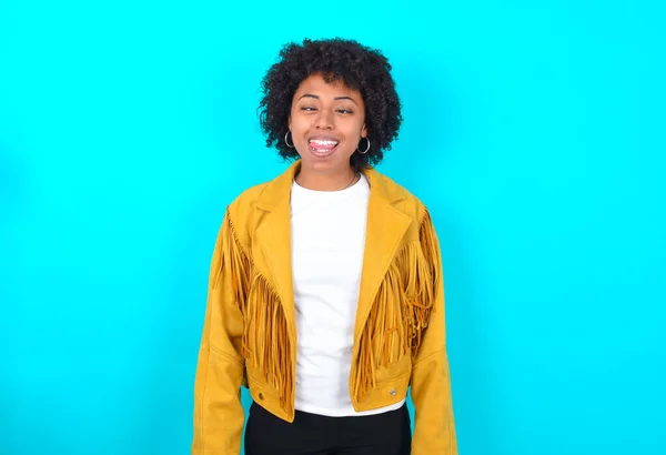 Funny Young African American woman wearing yellow fringe jacket over blue background makes grimace and crosses eyes plays fool has fun alone sticks out tongue.