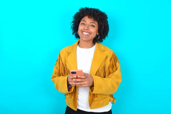 Młoda Afroamerykanka Ubrana Żółtą Frędzlową Kurtkę Niebieskim Tle Korzystanie Telefonu — Zdjęcie stockowe