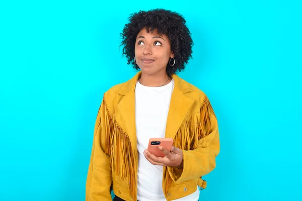 Młoda Afroamerykanka Żółtej Kurtce Frędzlami Niebieskim Tle Trzyma Telefon Komórkowy — Zdjęcie stockowe