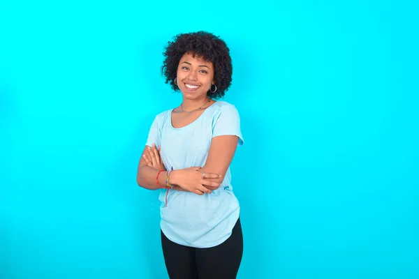 Portrait of African American woman with afro hairstyle wearing blue T-shirt over blue background standing with folded arms and smiling
