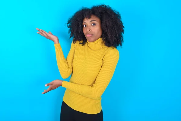 African American woman wearing yellow turtleneck over blue background pointing aside with both hands showing something strange and saying: I don\'t know what is this. Advertisement concept.
