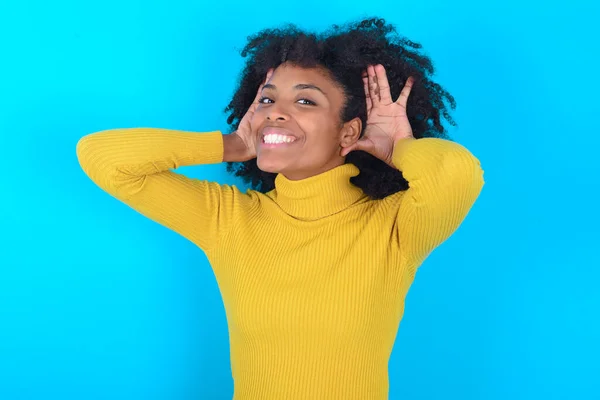 African American woman wearing yellow turtleneck over blue background Trying to hear both hands on ear gesture, curious for gossip. Hearing problem, deaf
