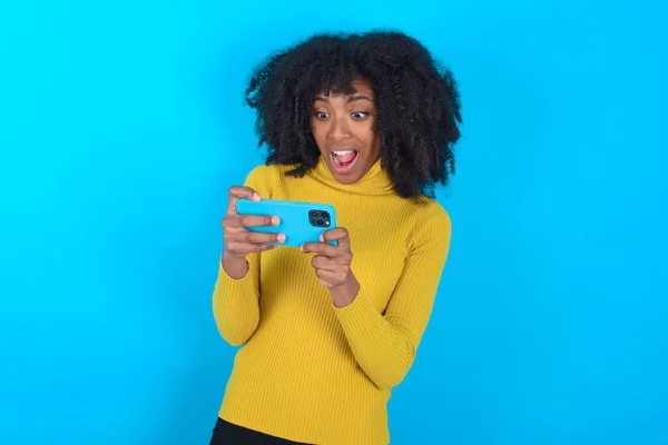 Portrait of an excited African American woman wearing yellow turtleneck over blue background playing games on mobile phone.
