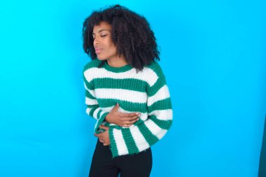 Mavi arka planda çizgili süveter giyen Afro-Amerikalı kadının midesi ağrıyordu.