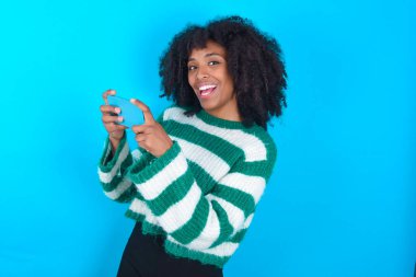 İyi bağımlı Afro-Amerikalı kadın mavi arka planda çizgili süveter giyiyor. Ağ oyunu oynuyor.