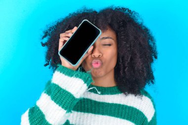 Mavi arka planda çizgili süveter giyen sevimli Afro-Amerikalı kadın dudaklarını büzmüş modern bir aletle gözlerini kapatıyor.