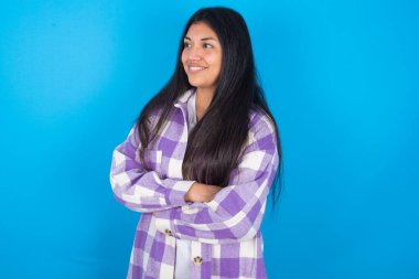 Mavi arka plan üzerinde ekose gömlek giyen genç Latin kadın çapraz eller fotokopi alanı gibi görünüyor.