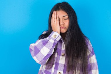 Mavi arka planda ekose gömlek giyen yorgun, yorgun, genç Latin bir kadın uykulu bir ifadeye sahip, kasvetli bir görünüme sahip, yüzü elle kaplanmış, gözleri kapalı, yorgunluktan soluk soluğa kalmış, partiden sonra bitkinlik