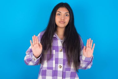 Mavi arka plan üzerine ekose gömlek giyen genç, ciddi Latin kadın avuç içlerini kameraya çekiyor, jestleri durduruyor, duygularını kontrol etmesini ve gergin olmamasını istiyor.