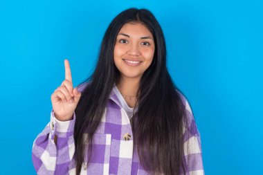 Mavi arka planda ekose gömlek giyen genç Latin kadın bir numaralı parmağı gösterip işaret ederken kendinden emin ve mutlu bir şekilde gülümsüyor..