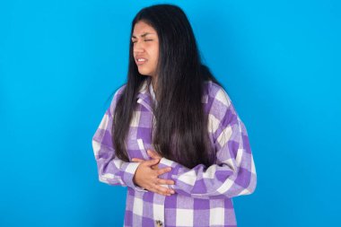 Mavi arka planda ekose gömlekli genç Latin kadın mide bulantısı, acı verici bir hastalık yüzünden elleri karnında. Ache kavramı.