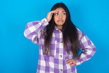 Mavi arka planda ekose gömlekli genç Latin bir kadın endişeli ve stresli bir şekilde elini alnından tutuyor..