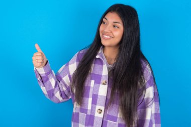 Mavi arka planda ekose gömlekli genç Latin bir kadın gururlu, gülümseyen, baş parmak kaldıran bir jest yapıyor. İyi iş.!