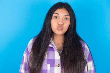 Mavi arka planda ekose gömlek giyen, dudaklarını büzen, kameraya bakan, insan yüz ifadeleri takınan hoş görünümlü genç Latin kadın.