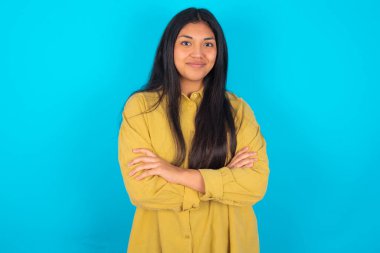 Kendine güveni tam, sakin, mavi arka planda sarı gömlek giyen Latin bir kadın kollarını kavuşturmuş duruyor. Profesyonel titreşimlerin kendine güvenen pozlarda durduğunu gösterir.