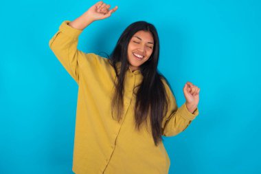 Mavi arka planda sarı tişört giyen neşeli Latin bir kadının fotoğrafı eğlenceli ve tasasız bir şekilde dans ediyor ve mükemmel bir ruh hali içinde hareket ediyor. Disko partisinde boş zaman geçirir