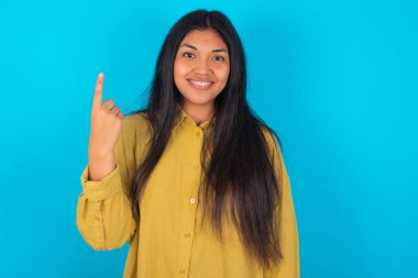 Mavi arka planda sarı tişörtlü genç Latin kadın gülümsüyor ve arkadaş canlısı görünüyor, bir numarayı gösteriyor ya da ilk eli önde, geri sayıyor.