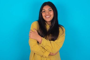 Mavi arka planda sarı tişört giyen hoş Latin bir kadın kendi vücudunu kucaklıyor, rahat pozlar veriyor. Şefkat ve kendine saygı kavramı