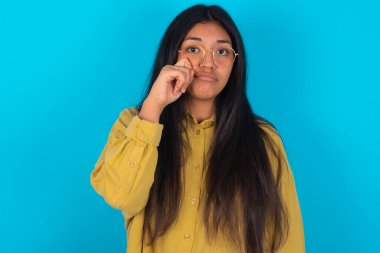 Hüsrana uğramış, mavi arka planda sarı gömlek giyen genç Latin kadın gözyaşlarını kasvetli bir ifadeyle tutuyordu. Olumsuz duygu