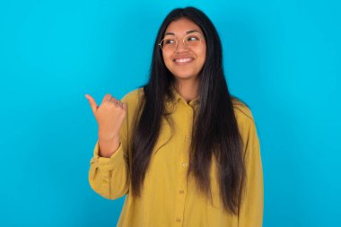 Mavi arka plan üzerinde sarı tişört giyen genç Latin kadın buradan işaret ediyor ve reklamını gösteriyor.