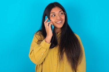 Mavi arka planda sarı tişörtlü mutlu, genç, Latin bir kadın. Hoş bir telefon konuşması yapıyor ve yan gelip bakıyor. Cep telefonuyla konuşurken güzel havasında ve gülümsüyor.