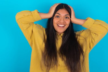 Mavi arka plan üzerine sarı tişört giyen neşeli, neşeli genç Latin kadın harika haberler aldıktan sonra ellerini havaya kaldırarak tepki veriyor..