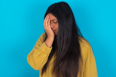 Mavi arka planda sarı tişört giyen genç Latin bir kadın. Ağlarken suratını ellerle kapatan üzgün bir ifade. Depresyon kavramı.