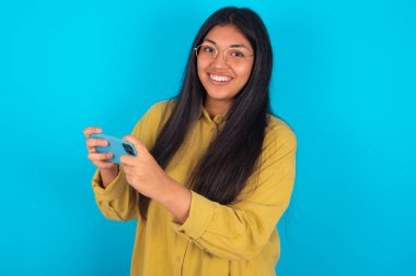 İyi bağımlı, neşeli, latin kadın. Mavi arka plan üzerinde sarı tişört giyiyor. Ağ oyunu oynuyor.