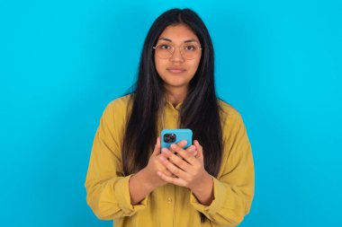 Mavi arka planda sarı tişörtlü, kendine güvenen genç bir kadının portresi. Telefonu iki eliyle tutuyor.