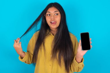 Mavi arka planda sarı tişört giyen hoş, genç bir Latin kadının fotoğrafı telefon ekranı saç kuyruğu olduğunu gösteriyor.