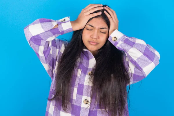 Mavi arka plan üzerine ekose gömlek giyen genç Latin kadın aşırı çalışmaktan dolayı şiddetli baş ağrısı ve stresten muzdarip. Depresyon ve acı kavramı.