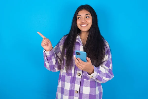 Wow!! excited young latin woman wearing plaid shirt over blue background showing mobile phone with open hand gesture